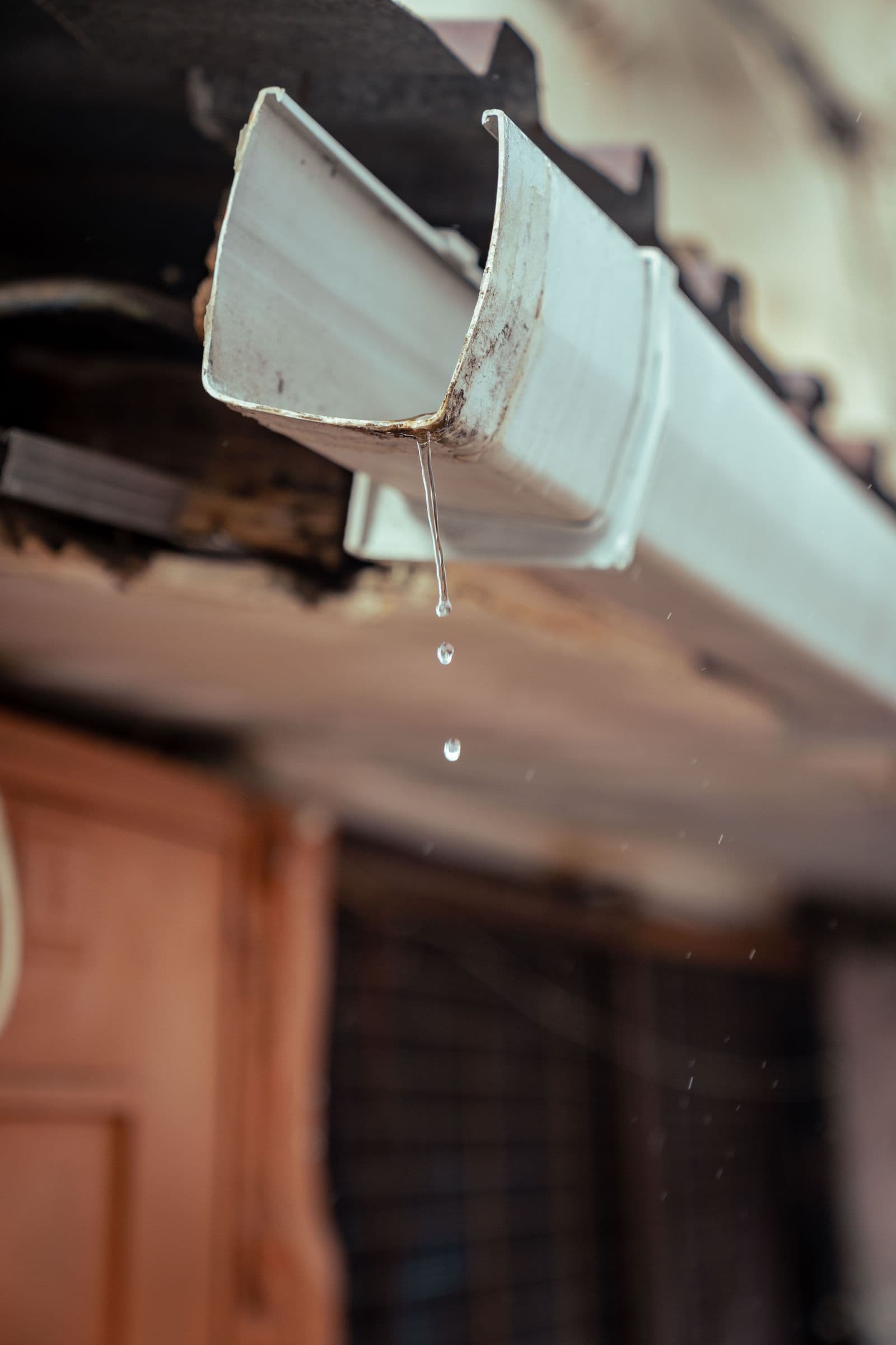 Weathered rain gutter along a roofline with water dripping from the end, the kind of clogged gutter that needs clearing before winter.