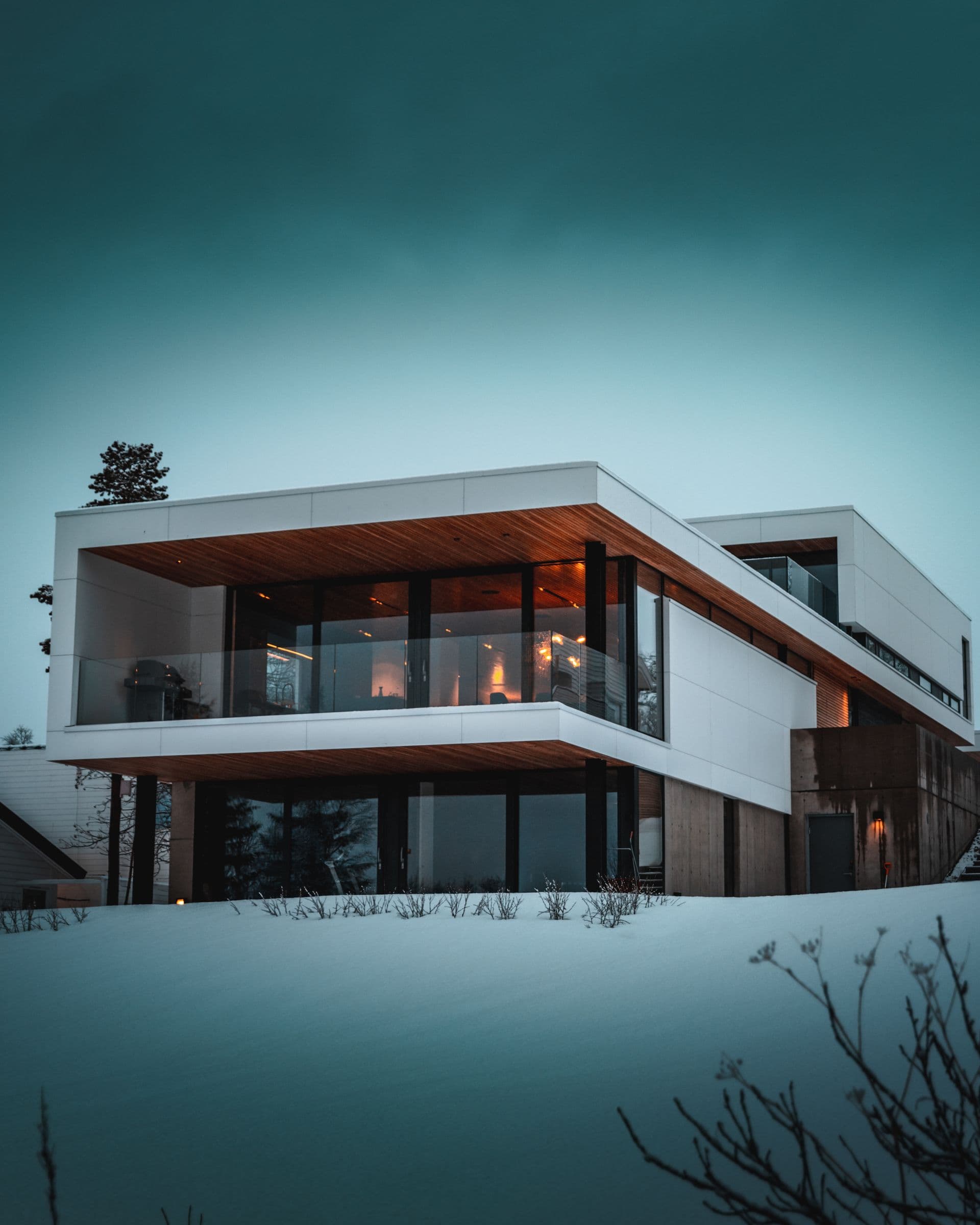 Modern glass-walled home lit from within at dusk in the snow, the kind of property kept clear year-round on a recurring maintenance plan.