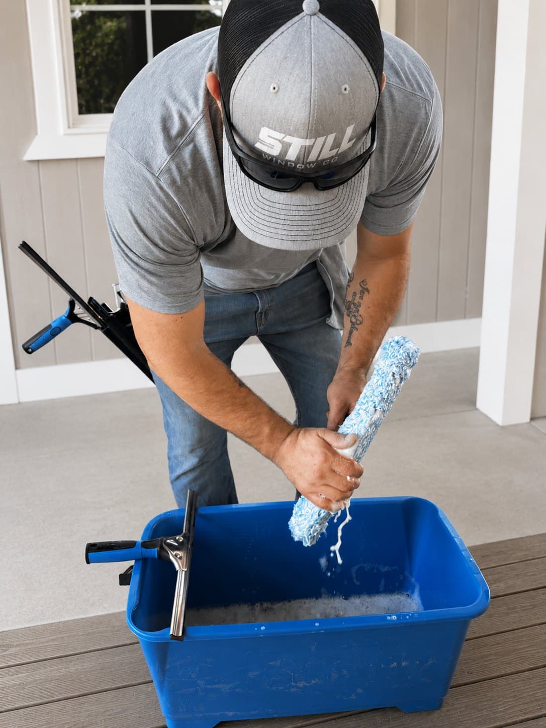 Still Window Co's owner wringing out a window-washing applicator over a bucket of soapy water, squeegees resting on the rim.