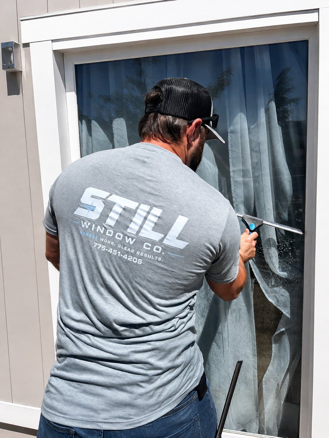 Still Window Co's owner squeegeeing a sliding glass door, wearing a branded Still Window Co shirt reading Honest work. Clear results.